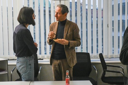 Two people talking in an office with blinds in the background.